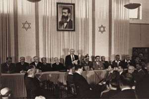 David Ben-Gurion (primul prim-ministru al Israelului), pronunţând public Declaraţia Statului Israel, la 14 mai 1948 (Tel Aviv, Israel), sub un portret mare al lui Theodor Herzl, fondatorul sionismului politic modern, în vechea clădire a Muzeului de Artă din Tel Aviv. (Sursa foto: Israel Ministry of Foreign Affairs, Autor: Rudi Weissenstein)